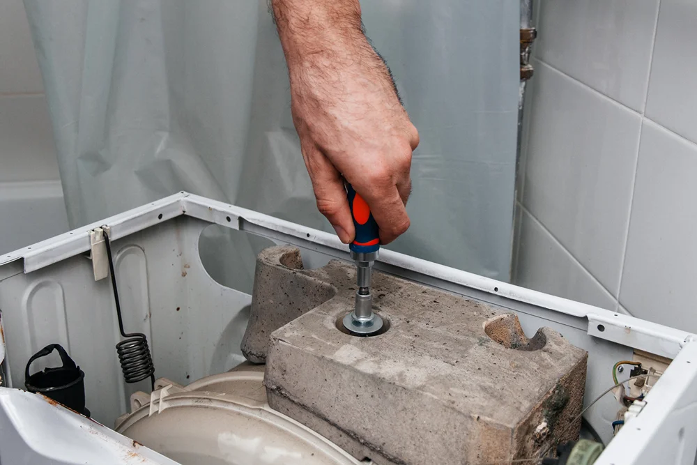 Bir el, çamaşır makinesinin içindeki beton ağırlığın vidasını elektrikli tornavidayla söküyor